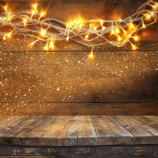 میز تخته ای چوبی در مقابل چراغ های گلدسته طلایی گرم کریسمس در زمینه چوبی روستایی تصویر فیلتر شده تمرکز انتخابی پوشش زرق و برق