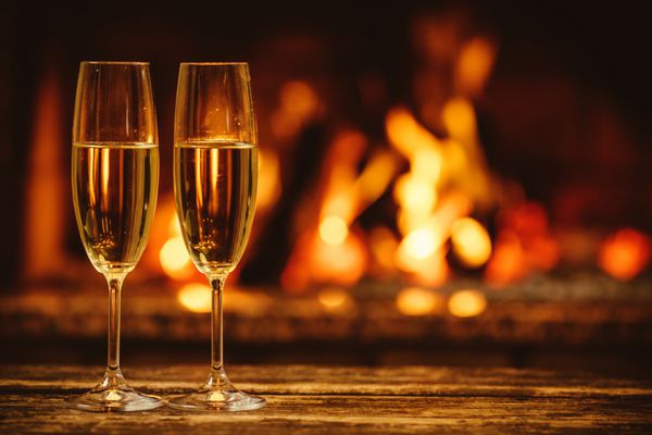 دو لیوان شامپاین درخشان جلوی شومینه گرم فضای جادویی دنج و آرام در خانه کلبه ای در کنار آتش مفهوم تعطیلات راحت پس زمینه زیبا با درخشان