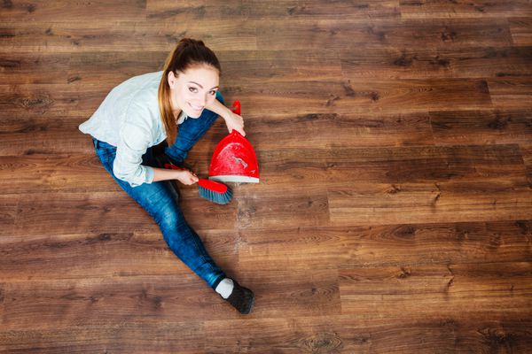 مفهوم پاکسازی کار خانه زن نظافتچی در حال جارو کردن کف چوبی با جارو کوچک قرمز رنگ و گردگیر و نمای با زاویه بالا