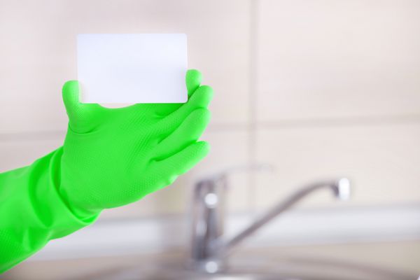 نزدیک دست نظافتچی خانه با دستکش ایمنی که کارت ویزیت خالی را در مقابل سینک آشپزخانه و کاشی‌ها در پس‌زمینه نشان می‌دهد