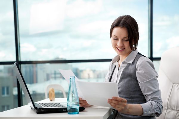 زن تاجر زیبا در حالی که در دفتر کارش روی کامپیوتر کار می کرد به کاغذهایی که در آغوش گرفته نگاه می کند