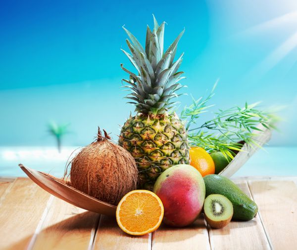 میوه های تازه در ساحل در یک عرشه در مقابل یک جزیره با یک نخل انواع میوه های استوایی پرتقال آناناس یا آناناس لیموترش انبه و آووکادو
