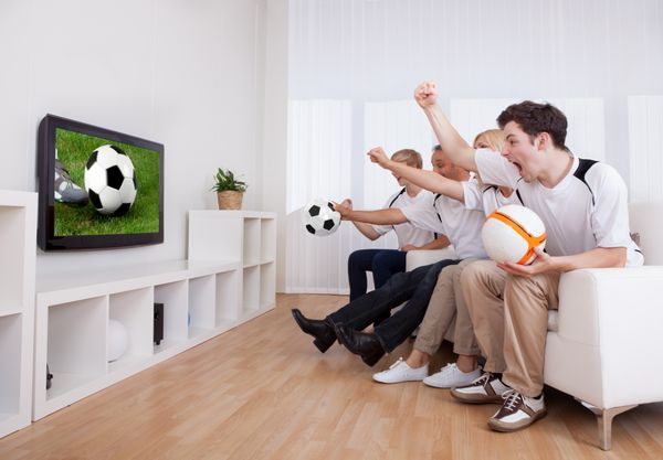 خانواده شاد در حال تماشای تلویزیون در حالی که طرف خانه خود را در یک مسابقه ورزشی تشویق می کنند
