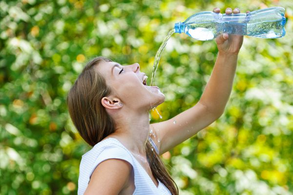 زن جوان زیبا در برابر سبز پارک تابستانی از بطری آب می نوشد