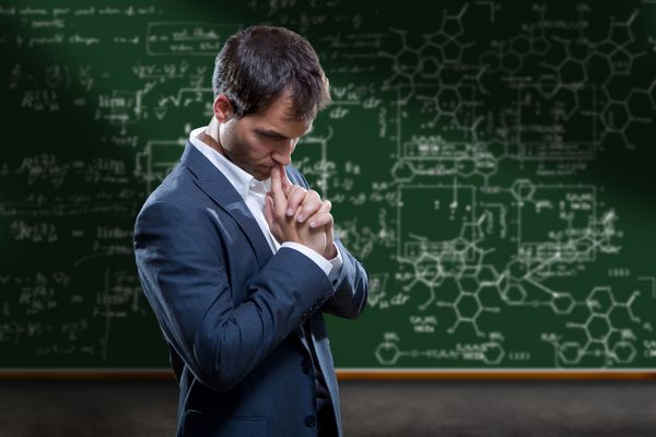 یک معلم دانشمند یا استاد کالج که در مقابل یک تخته سیاه با معادلات پیچیده ریاضی ایستاده و فکر می کند