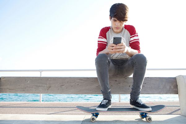 پسر نوجوان جذاب و متفکری که با اسکیت بورد استراحت می کند و روی یک نیمکت کنار دریا می نشیند گوشی هوشمندی را برای شبکه در یک روز آفتابی در فضای باز در دست گرفته و از آن استفاده می کند