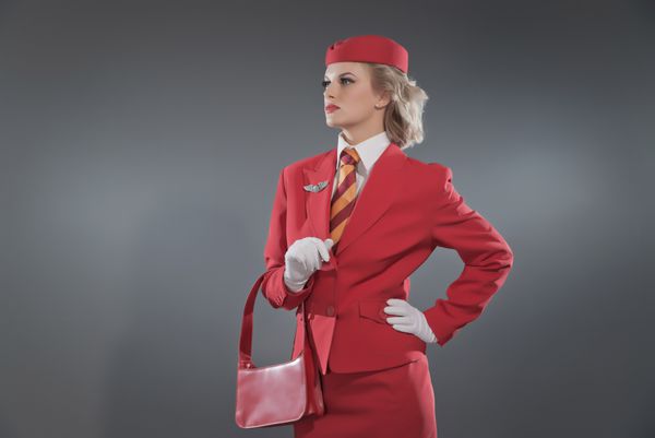 مهماندار رترو با کت و شلوار قرمز با کراوات راه راه و کلاه در دست گرفتن کیف چرمی قرمز استودیو در برابر خاکستری