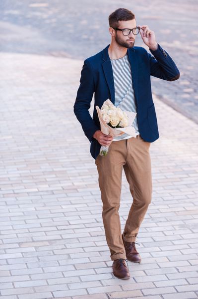 بهترین گل ها برای او مرد جوان خوش تیپ تمام قد با لباس هوشمند که دسته گل در دست دارد و هنگام راه رفتن در خیابان عینک خود را تنظیم می کند