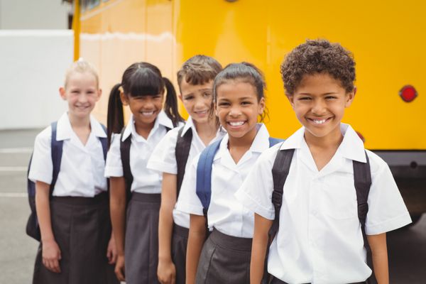 بچه های مدرسه ای زیبا که در کنار اتوبوس مدرسه بیرون از مدرسه ابتدایی به دوربین لبخند می زنند