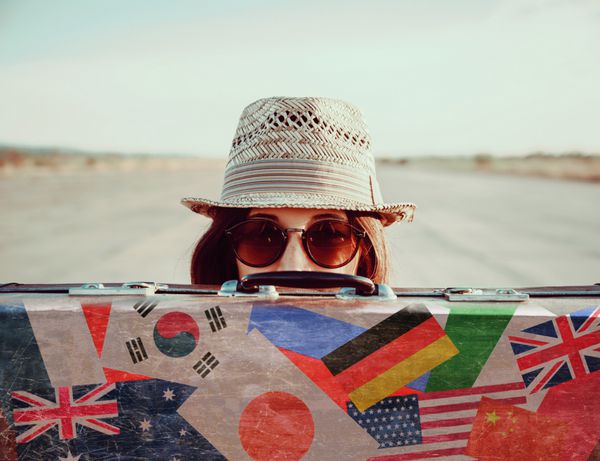 دختر هیپستر با کلاه و عینک آفتابی از چمدان قدیمی به نظر می رسد چمدان با تمبر پرچم کشورهای مختلف