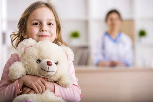 دختر بچه با خرس عروسکی به دوربین نگاه می کند دکتر زن در پس زمینه