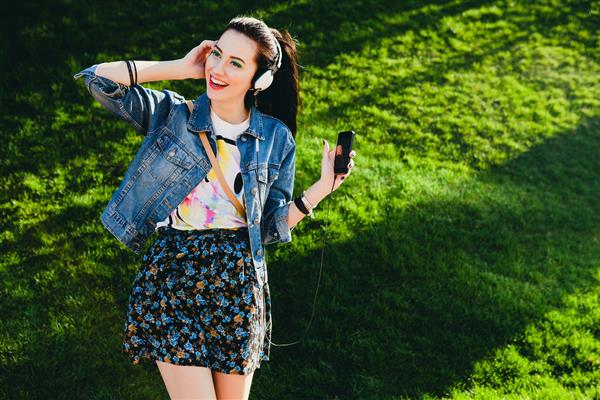 دختر جوان هیپستر شیک و زیبا در حال گوش دادن به موسیقی تلفن همراه هدفون لذت بردن لباس جین خندان شاد لوازم جانبی جالب سبک قدیمی سرگرمی خندیدن پارک