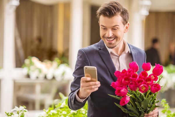 مرد خوش تیپی که دسته گل لاله در دست دارد و به تلفن نگاه می کند قرار عاشقانه
