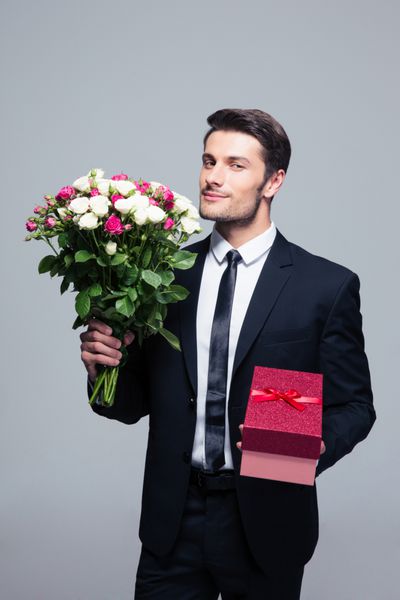 تاجر خوش تیپ که گل و جعبه هدیه را روی پس زمینه خاکستری نگه می دارد نگاه کردن به دوربین