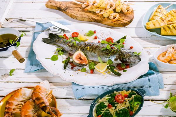 ماهی کامل کبابی تزیین شده روی بشقاب سفید روی میز پیک نیک سفید روستایی احاطه شده با ظروف سبزیجات و غذاهای دریایی و دستمال های آبی رنگ