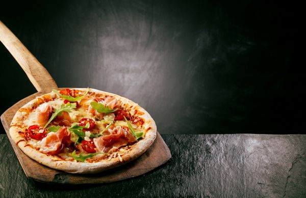 ژامبون گوجه فرنگی و آروگولا یا پیتزای غیره که در پیتزا فروشی یا رستوران روی یک تخته چوبی دسته بلند روی پیشخوانی روستایی با کپی پشت آن سرو می شود