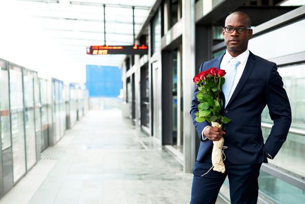 تاجر جوان آفریقایی آمریکایی در مترو ایستاده و دسته گل رز را در دست دارد و به دوربین نگاه می کند