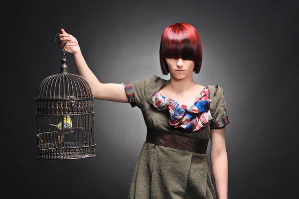 یک دختر جوان زیبا که یک قفس پرنده را با پرنده ای درون آن در پس زمینه ای تاریک نگه داشته است