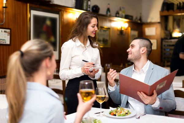 زوج زیبایی که در رستورانی غذا می خورند در حالی که پیشخدمت خوشحال در حال گرفتن سفارش آنهاست