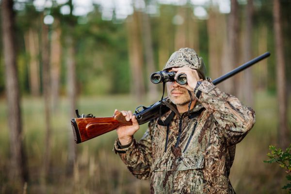 مرد شکارچی با تفنگ که از طریق دوربین دوچشمی در جنگل نگاه می کند