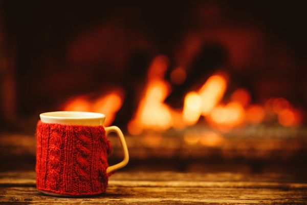 فنجان نوشیدنی در مقابل شومینه گرم مفهوم تعطیلات کریسمس لیوان با دستکش بافتنی قرمز که در نزدیکی آتش ایستاده است فضای جادویی دنج و آرام در یک کلبه