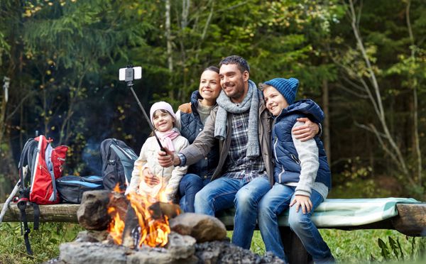 کمپینگ مسافرت گردشگری پیاده روی و مفهوم مردم - خانواده شاد روی نیمکت نشسته و با گوشی هوشمند روی چوب سلفی در آتش کمپ در جنگل عکس می گیرند