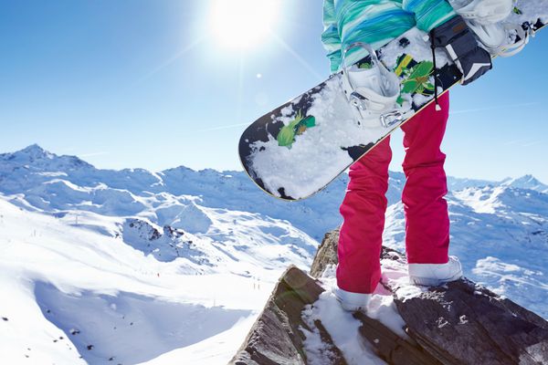 نمای عقب از نزدیک اسنوبرد زن با کلاه ایمنی ژاکت آبی دستکش خاکستری و شلوار صورتی ایستاده با اسنوبرد در یک دست و لذت بردن از منظره کوهستانی کوهستانی - مفهوم ورزش های زمستانی