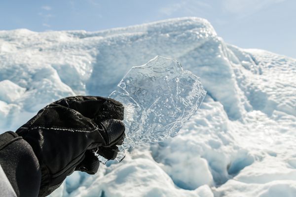 دست با دستکش که یک تکه یخ شفاف با پس زمینه برف و یخ در دست دارد