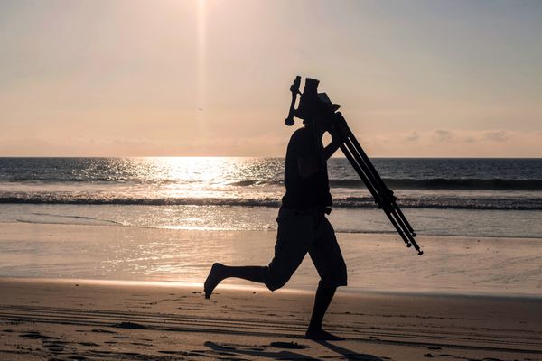 سیلوئت مرد حرفه ای عکاس که با سه پایه اش در ساحل گالاپاگوس آمریکای جنوبی می دود
