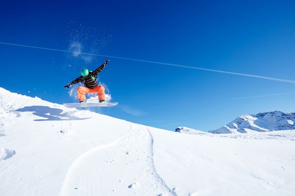 اسنوبردباز مرد با کلاه ایمنی سبز ژاکت مشکی و شلوار نارنجی در حال پریدن در مقابل آسمان آبی در پیست پیست اسکی - مفهوم ورزش های زمستانی