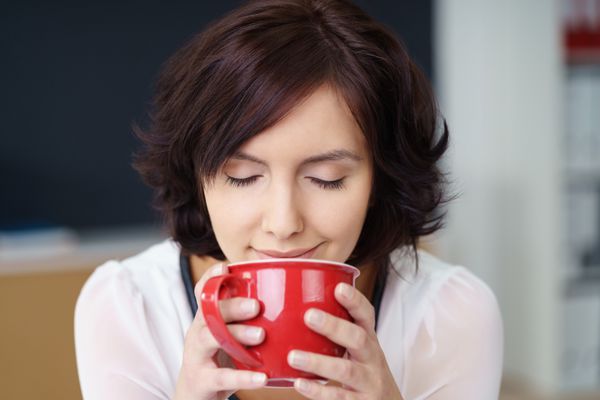 از نزدیک زن اداری جوان و زیبا که عطر نوشیدنی قهوه خود را در فنجان قرمز با چشمان بسته استشمام می کند