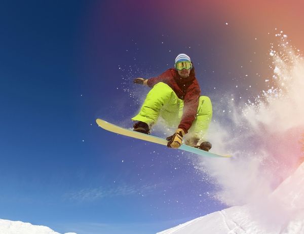 اسنوبورد پرش یک دست خود را روی اسنوبرد در کوهستانی در پیست اسکی در پس زمینه آسمان آبی نگه می دارد