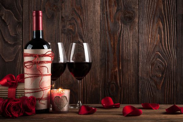 بطری قرمز دو لیوان جعبه هدیه شمع و گل رز قرمز روی زمینه چوبی تیره