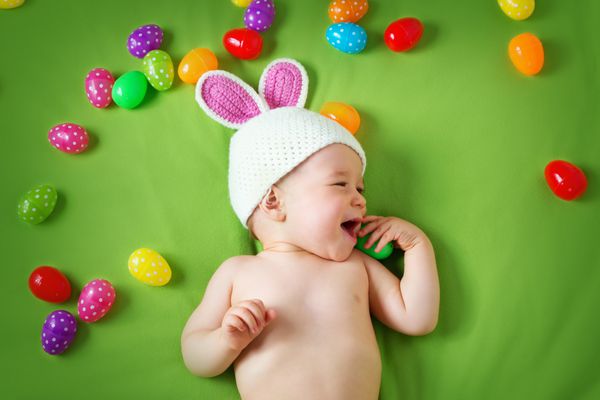 پسر بچه با کلاه اسم حیوان دست اموز روی پتوی سبز با تخم مرغ های عید پاک دراز کشیده است