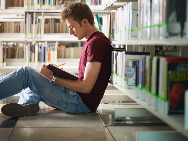 دانشجوی مرد در کتابخانه روی زمین نشسته و مشغول خواندن کتاب است شکل افقی نمای جانبی طول سه چهارم کپی sp