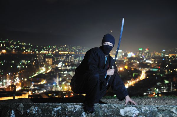 قاتل نینجا شمشیر جنگی قدیمی سامورایی کاتانا را در شب با چراغ های شهر در پس زمینه نگه می دارد