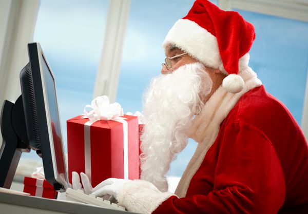 پرتره بابا نوئل در مقابل مانیتور کامپیوتر با هدایا