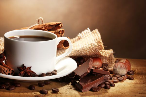 فنجان قهوه و دانه ها چوب دارچین آجیل و شکلات روی میز چوبی در زمینه قهوه ای