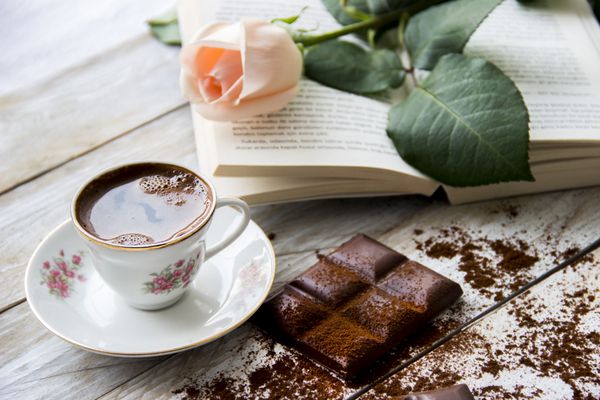 قهوه ترک با گل رز هلویی رنگ تکه های شکلات و کتاب روی میز چوبی
