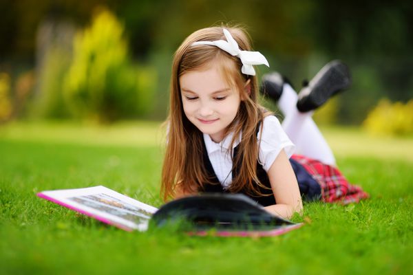 دختر کوچولوی شایان ستایشی که روی چمن دراز کشیده و کتاب می خواند