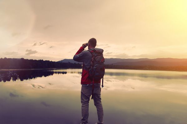مرد جوان با یک کوله پشتی در نزدیکی دریاچه ایستاده و از طریق دوربین دوچشمی نگاه می کند رنگ قدیمی