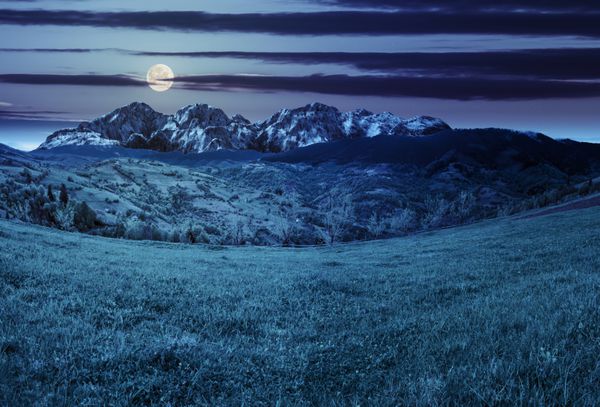 منظره تابستانی ترکیبی با کوه های سه بعدی روستایی در دامنه تپه چمنزار کشاورزی در مقابل یک رشته کوه مرتفع در شب در نور ماه کامل