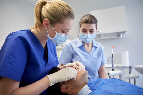 مفهوم مردم پزشکی دهان و دندان و مراقبت از دندان - دندانپزشک زن با دستیار در حال بررسی دندان های انسداد دندان بیمار مرد در مطب کلینیک دندانپزشکی