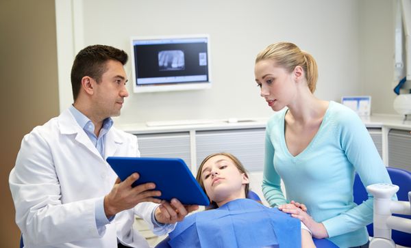مردم پزشکی دندان‌شناسی فناوری و مفهوم مراقبت بهداشتی - دندانپزشک مرد در حال نشان دادن رایانه تبلت به دختر بیمار و مادرش در مطب کلینیک دندانپزشکی