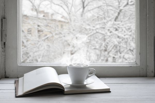 طبیعت بی جان زمستانی دنج فنجان قهوه و کتاب باز شده روی طاقچه پنجره قدیمی در برابر منظره برفی از بیرون