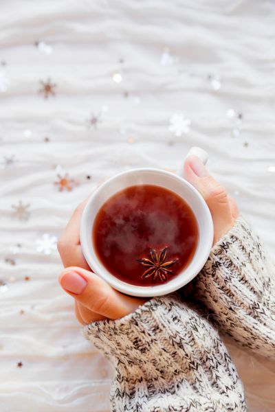 زن یک فنجان چای با ستاره انیسون در دست دارد پس زمینه پارچه زمستانی با دانه های برف نقره ای درخشان