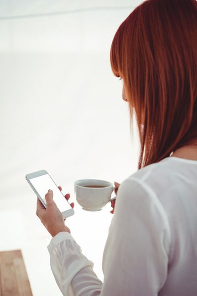 نمای پشت زنی با موهای قرمز با استفاده از گوشی هوشمند و در دست گرفتن فنجان قهوه