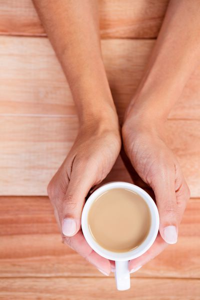 دست های زن که فنجان قهوه را روی میز چوبی گرفته اند