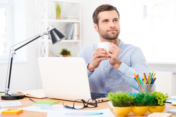 یک فنجان قهوه برای ایده های جدید مرد جوان خوش تیپ با پیراهن که یک فنجان قهوه در دست دارد و در حالی که در محل کار خود نشسته است به دور نگاه می کند