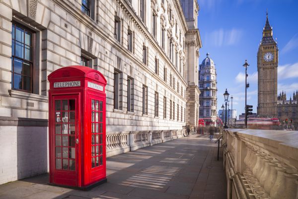 جعبه تلفن سنتی قرمز بریتانیایی با اتوبوس بزرگ و دو طبقه در پس‌زمینه در یک بعد از ظهر آفتابی با آسمان آبی و ابرها - لندن انگلستان